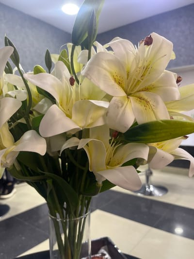 This image, captured in Talin, Armenia, presents a close-up perspective of an artificial floral arrangement. The primary subject is a dense bouquet of white lily-type flowers with prominent yellow centers and red-brown anthers, displayed in a clear cylindrical glass vase. Several flowers show minor brown discoloration or marks on their petals. Green stems and leaves are visible, intermingled with the blooms and a few closed flower buds. The vase rests on a dark, reflective surface, which itself is situated on a patterned floor or tabletop consisting of alternating rectangular dark grey/black and light beige/cream tiles arranged in parallel stripes. In the blurred background to the right, the polished metallic base of a chair or stool is visible, reflecting light. The upper background features a plain light grey wall, and a bright, circular light fixture on the ceiling or upper wall emits a strong white light and a visible lens flare. No individuals or specific actions are depicted.