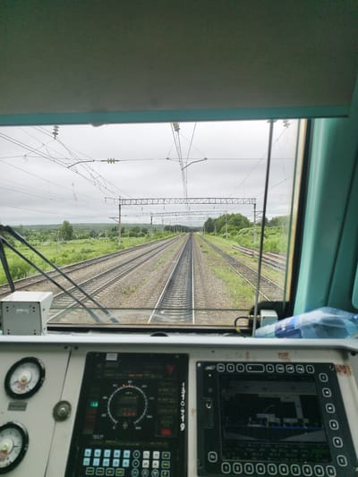 The image is taken from the perspective of a train driver inside a locomotive cab, looking out through the windshield at a set of railway tracks receding into the distance. The tracks are double-gauged and appear to be on an overcast day. Lush green vegetation lines the sides of the tracks, with trees and some small structures visible in the background, suggesting a rural or semi-rural setting. Overhead, a complex network of power lines and supporting structures for an electric railway is visible against the cloudy sky.

In the foreground, the interior of the locomotive cab is partially visible. There are several control panels with buttons, dials, and a digital display screen. Two circular gauges with needles are prominent on the left side. A white box-like device is mounted on the windshield support. A blue plastic bottle is visible on the right side of the cab.

The location is identified as Daktui, Russia. The scene depicts a train on an operational railway line, likely in transit, with no people or specific actions immediately evident, beyond the implied operation of the train.
