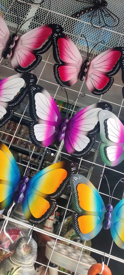 A close-up shows an arrangement of several colorful metal butterflies on a white wire rack. The butterflies come in shades of pink, purple, and rainbow hues. A metal dragonfly decoration is visible in the upper left corner. The wire rack also contains other items, including a ceramic bowl and miscellaneous objects wrapped in plastic, indicating a possible retail or display setting. The location is Tres de Mayo, Mexico, though there is no locational data in this image.