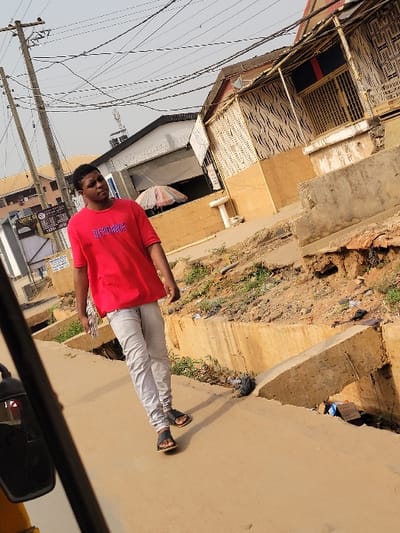 The scene depicts an urban street in Puerto López, Colombia, under an overcast sky. A male individual, dressed in a red short-sleeved t-shirt with light-colored text, light grey pants, and black sandals, walks on a concrete pathway towards the upper left. He holds a folded white object in his left hand. To the right of the pathway, a concrete drainage channel contains exposed soil, sparse weeds, a black plastic bag, and a cardboard box. Multiple buildings form the background; the prominent structure on the right has a light brown lower facade and an upper section with a black and white wavy pattern, featuring a balcony with metal grates. Behind the walking individual, a building with a dark facade is visible, in front of which an open white umbrella is positioned. Multiple utility poles line the left side, supporting a dense array of overhead power lines. A portion of a vehicle's side mirror is visible in the lower-left foreground.