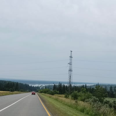 The image captures a view from a road, likely in or near Нечкино, Russia, under an overcast sky. The foreground features a two-lane asphalt road with a white line dividing the lanes and a yellow line marking the right edge of the road. A red car is visible driving away from the viewer on the right lane. The roadside is bordered by green grass and tall, dry vegetation.
In the middle ground, a tall metal electricity pylon stands prominently to the right of the road. Several power lines stretch across the frame, disappearing into the hazy distance. Beyond the roadside, a dense evergreen forest lines the landscape, sloping downwards towards a body of water, possibly a river or lake, which is visible in the far distance. Small clusters of buildings and a white church spire are visible amidst the trees on the far bank of the water. The overall atmosphere is serene and natural, with the presence of infrastructure suggesting a rural or semi-rural setting.