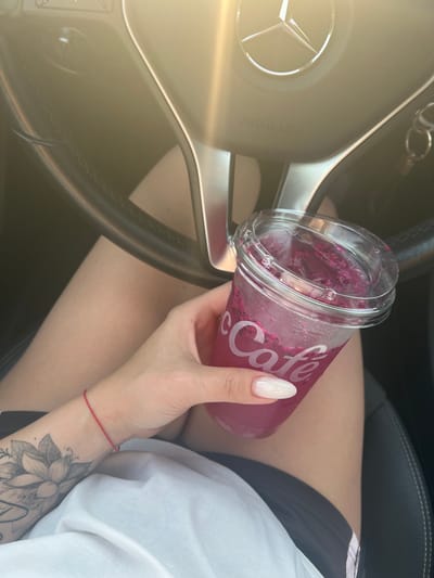 A person is holding a pink, iced beverage in a clear plastic cup with the "McCafé" logo visible on it. The cup contains pieces of fruit. The person's hand has long, white fingernails and a red string bracelet on their wrist. They also have a tattoo of a flower on their forearm. They are wearing a white top and dark shorts. The person is seated in a car, indicated by the steering wheel with a Mercedes-Benz logo and the word "AIRBAG" printed on it, and black leather interior. The scene appears to be outdoors, with sunlight creating a glare on the steering wheel. The location is a Mercedes-Benz vehicle, likely in Heinsberg, Germany.