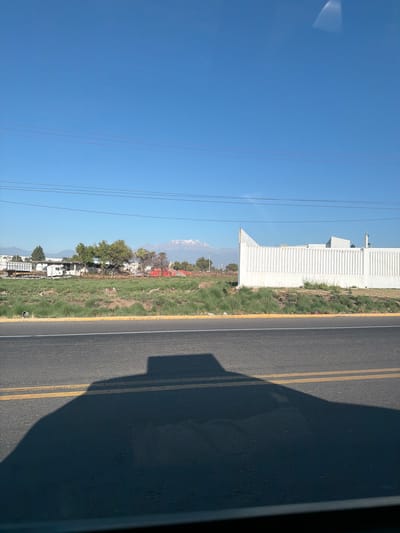 This image depicts a roadside scene in Santa María Zacatepec, Mexico, under a clear blue sky. In the foreground, the dark, elongated shadow of a vehicle occupies the lower half of an asphalt road, which features a solid white line at its upper edge and a double solid yellow line further down. Beyond the road, a strip of unmaintained grassy land with scattered dirt patches extends horizontally. To the right, a tall, white wall with vertical ridging encloses a building, partially visible above the wall. To the left of this wall, an open lot contains sparse grass, several trees (some with green foliage, others appearing dry), and stacks of reddish-orange objects, possibly construction materials. A white truck with a flatbed trailer is parked in this area, adjacent to what appears to be an open-sided shelter. Multiple utility power lines traverse the upper midground. The distant background reveals a prominent snow-capped mountain peak, flanked by other mountainous terrain, all set against a clear, bright blue sky. No individuals are visible.