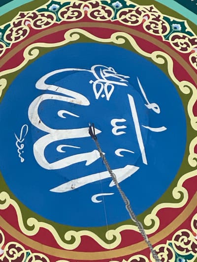 This image depicts a segment of a richly decorated ceiling or dome interior, located in Mns Asan, Indonesia. A central circular panel, painted royal blue, features prominent white Arabic calligraphy; the word "Allah" (الله) is clearly visible, with additional smaller script present to the left. A silver-colored metal chain, secured by a dark clip, descends diagonally from the approximate center of the blue panel. A thin, translucent wire or string is also visible adjacent to the chain. The blue panel is encircled by multiple concentric decorative bands: an immediate inner wavy light yellow border, followed by a wider red band, a second wavy light yellow border, a dark green band, and an outermost light green band adorned with intricate patterns featuring small blue and dark red floral or geometric motifs. No individuals are present in the scene.