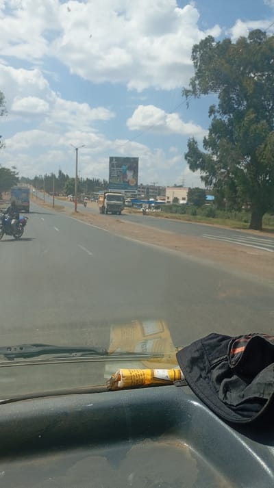 A view from inside a vehicle shows a road with several vehicles, including a motorcycle and a truck. A large billboard is visible in the distance. The sky is partly cloudy with bright sunlight. The vehicle's dashboard has a rolled-up tube and a black hat on it. The road appears to be a highway with lanes going in both directions. Some buildings are also visible along the roadside.
