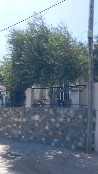 This is a horizontal, slightly blurry, eye-level, outdoor, daytime photograph taken from a moving vehicle. The scene appears to be a coastal area in the city of Ηρακλής, Greece. In the foreground, there is a rough stone wall constructed from irregularly shaped, multi-colored stones. Beyond the wall, a building with white shutters is visible. An old, gnarled tree with lush green foliage partially obscures the building. There are several chairs on a patio area in front of the building. A utility pole with a sign is positioned on the right side of the frame. The sky is clear and blue.