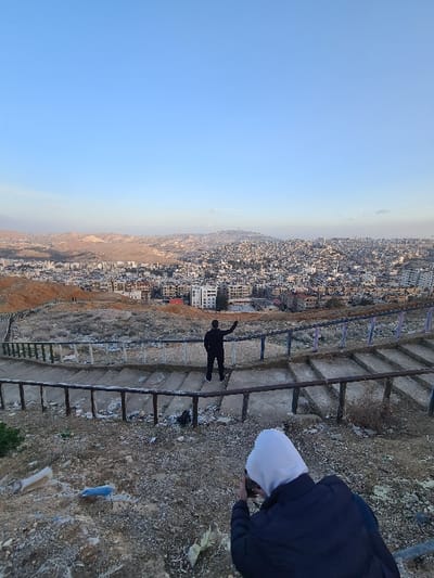 The image presents an elevated view overlooking the city of Qudsaiya, Syria, under a clear blue sky. In the immediate foreground, a person wearing a dark jacket with a white hood is observed from behind, holding a camera or smartphone in a shooting position. Ahead of this individual, a second person in dark clothing and light shoes stands on a descending concrete staircase, also viewed from the rear, facing the distant urban panorama. This second individual's right arm is raised and extended, with fingers splayed. The hillside in the foreground and around the staircase consists of dry, rocky earth with sparse vegetation and scattered debris. The staircase itself features worn railings. In the background, the dense urban area of Qudsaiya stretches across valleys and hills, characterized by numerous light-colored, multi-story buildings. Beyond the city, arid, light brown hills extend towards the horizon.