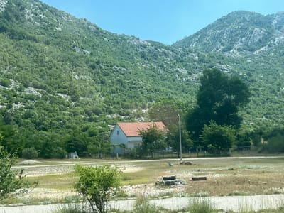 The image depicts a rural landscape in the vicinity of Dragalj, Montenegro. In the foreground, a dry, grassy field with scattered debris and vegetation extends towards a paved road. A small, white house with a red tile roof is situated in the mid-ground, surrounded by trees and a fence. Behind the house, a steep, densely forested mountain rises sharply, with rocky outcrops visible through the greenery. The sky above is clear and blue. The scene appears to be captured during daylight hours.
