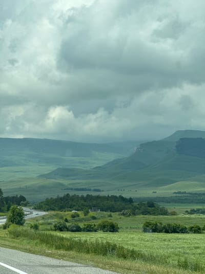 The image shows a wide, green valley with rolling hills and mountains in the distance under a cloudy sky. A road with a few cars is visible in the mid-ground, winding through fields and trees. The foreground is dominated by a grassy field and the edge of a paved road. The scene appears to be a rural landscape, possibly in the vicinity of Кубина, Russia, characterized by lush vegetation and a somewhat overcast atmosphere.