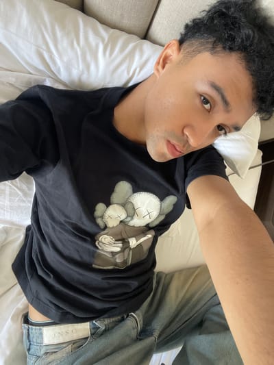 The image depicts a young adult male lying on his back, positioned diagonally across a bed or sofa, reportedly in Kayuambon, Indonesia, though no specific visual cues within the image confirm this geographical location. He is taking a selfie, with his right arm extended towards the camera, looking directly into the lens with a neutral expression.

The individual has a light-to-medium skin tone, dark, curly hair with faded sides, and dark eyes. He is wearing a black, short-sleeved t-shirt featuring a graphic design of multiple figures resembling KAWS Companions, characterized by "X" marks for eyes. One larger figure appears to be holding a smaller one; the colors of the graphic are muted greys, beige, and white. Below the t-shirt, light blue denim jeans are visible, with a white waistband bearing the label "JNSO" (or similar, partially obscured) and a thin light blue line showing above the jeans.

The subject is lying on white bedding, including a white pillow supporting his head. A light-colored, textured headboard or sofa backrest is visible in the background, behind his head. The lighting is soft and even, suggesting an indoor environment.