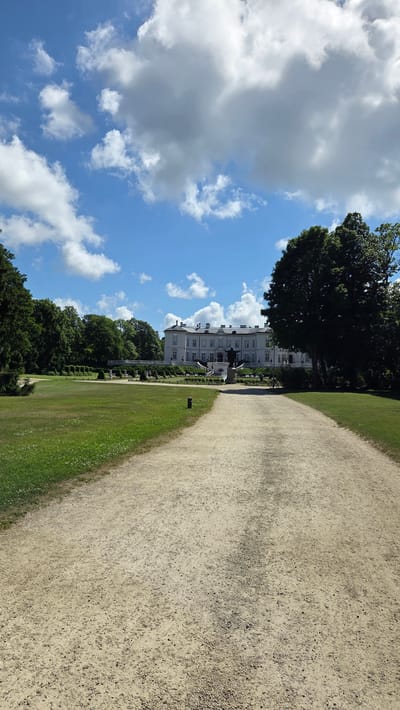 The image depicts a wide, gravel pathway leading towards a large, white manor-style building with a long facade and multiple windows. The building is situated amidst lush green grounds, with manicured hedges and scattered trees. A prominent dark-colored statue stands on a raised platform in front of the building, accessible by a set of stairs. The sky above is a clear blue, dotted with fluffy white clouds. The scene appears to be a well-maintained park or estate.

This location is the Palanga Amber Museum, also known as Tiškevičiai Palace, in Palanga, Lithuania.