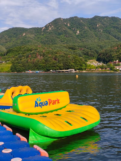 The image shows a vibrant yellow and green inflatable water park structure floating on a dark body of water. The inflatable has the words "Aqua Park" written in blue on its surface. In the background, a lush green mountain range rises under a bright blue sky with scattered clouds. The shoreline is lined with trees and some buildings, suggesting a recreational area. The scene appears to be located at the Chuncheon Aqua Park in Chuncheon-si, South Korea, a popular destination for water activities. There are no people visible in the immediate foreground of the image. The water is slightly rippled, and the inflatable is casting a reflection on its surface. The overall impression is one of a sunny day at an outdoor water amusement facility.