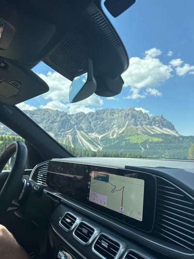 An interior shot from the passenger seat of a vehicle captures a scenic mountain landscape through the windshield under a partly cloudy blue sky. The dashboard is visible, featuring a large infotainment screen displaying a navigation map with directions to Landsteinerstraße 29. The steering wheel and air vents are also prominent. The mountain range in the background is rugged and appears to be in the Dolomites region of Italy, likely near San Martin de Tor - San Martino in Badia - St. Martin in Thurn, given the context. The scene suggests a road trip through a mountainous area.