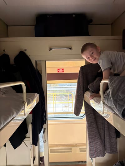 The image, oriented sideways, shows the interior of a train compartment, likely a sleeper car. A young boy with light hair, wearing a light grey long-sleeved shirt, is looking directly at the camera with a gentle smile. He is positioned near what appears to be a lower bunk or seat, with his head and upper torso visible.

The compartment features functional elements characteristic of a train. A white metal bed frame, draped with grey and white blankets or duvets, is prominent. A dark, houndstooth-patterned blanket or jacket is visible on a lower sleeping area. Various dark clothing items and a black bag are also present within the confines of the cabin. Silver grab handles are mounted on the light-colored walls.

Through a large window, an exterior view reveals bare trees or branches, suggesting a cool or wintery day, and some yellow structures in the distance. The lighting outside appears overcast, contributing to a muted daytime ambiance, while artificial light illuminates the interior.

A notable detail is a red sign visible through the window, partially obscured by the window frame. The Cyrillic text on the sign reads "ОКНО АВАРІЙНОГО ВЫХОДА," which translates to "EMERGENCY EXIT WINDOW." This text is a clear geographical indicator, consistent with the location context of Bobrov, Russia. The scene captures a quiet moment of travel or rest inside the train.