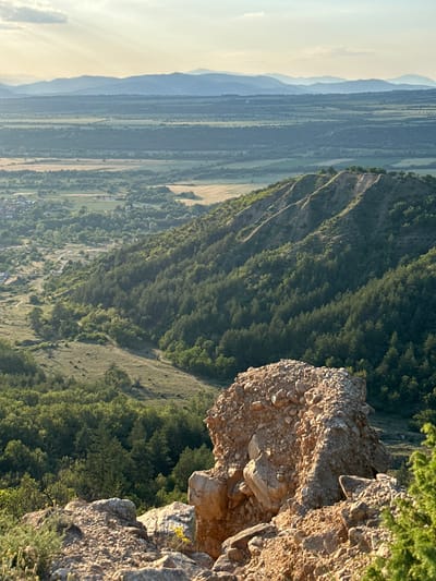 An elevated landscape view overlooks the Stob Pyramids natural rock formations and the surrounding area in Stob, Bulgaria. In the immediate foreground, a substantial, light brown rocky outcrop composed of many smaller stones and some larger boulders dominates the lower right, partially illuminated by warm, direct sunlight, casting subtle shadows. Sparse green vegetation, including small shrubs and grasses, grows around and on the rocks.
Beyond the foreground, the midground features steep, eroded hillsides covered predominantly with dense, dark green pine forests. Distinctive light brown ravines and gullies are visible on the slopes, indicative of the Stob Pyramids' unique geological formations. Winding dirt paths or unpaved roads traverse the gentler slopes and valleys between the forested areas. To the far left in the midground, a small cluster of houses with light-colored roofs, forming part of a village (likely Stob), is visible amidst green fields and trees.
The background presents an expansive, agricultural valley floor characterized by a patchwork of green fields, some appearing tilled or harvested, interspersed with sections of woodland. Beyond the valley, a long range of hazy blue-grey mountains stretches across the horizon under a pale sky, with diffused sunlight originating from the upper left. The overall lighting suggests either early morning or late afternoon, with a warm, soft glow. No people or animals are visible in the scene.