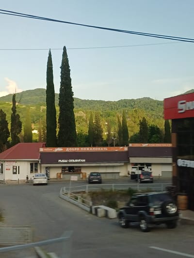 The image depicts a street scene in Kazachiy Brod, Russia. In the foreground, a dark-colored SUV is parked on the right side of the road, facing away from the camera. The road itself is paved and has white lane markings. On the left side of the road, there is a white building with a red roof and several windows. Above this building, an orange sign displays Russian text, including "ДОБРО ПОЖАЛОВАТЬ" (Welcome) and "РЫБЫ ОПЬЯНЕНЫ" (Drunken Fish). Further down the street, a white sedan is parked in front of another building. Behind these buildings, tall, slender trees, possibly cypresses, are visible, with lush green hills and mountains in the background under a clear sky. To the far right, a red building with white lettering is partially visible. Several cars are parked in the vicinity. The overall scene suggests a commercial area in a semi-rural or suburban setting.