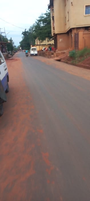 The image depicts a street scene in Nibo, Nigeria. A two-lane asphalt road, extensively covered with reddish-brown soil and dust, extends from the foreground towards the background under an overcast sky. In the immediate left foreground, the front quarter of a white and blue vehicle is partially visible. Further along the road, a white van is positioned on the right side, facing away from the viewer. A single individual, wearing a blue top, operates a motorcycle, traveling towards the viewer in the road's center-left.
On the right side of the road, a multi-story building, at least three stories tall, featuring a light beige facade and brown window frames, is prominent. Adjacent to the base of this building, a group of approximately seven individuals, dressed in various casual attire including bright colors, are congregated on a raised concrete platform, some seated. A white scooter is parked near this group. A concrete drainage ditch runs parallel to the road on the right. Dense green foliage is visible behind the buildings, extending into the background.