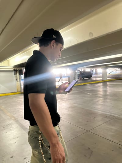 A male subject is depicted standing in a multi-story parking garage in Paradise, United States. He is viewed in profile, facing right, wearing a black baseball cap with a white logo on the side, a black short-sleeved polo shirt, and light-colored, possibly distressed, jeans. A gold-toned decorative chain, adorned with pearls and other elements, is attached to his left hip. He holds a smartphone in his right hand, directing his gaze downward towards its illuminated screen. Rings are visible on his fingers. A prominent lens flare extends horizontally across his torso. The environment features concrete ceiling beams and a concrete floor marked with yellow lines. In the background, a dark-colored SUV and several blurred human figures are visible, illuminated by bright ambient light from the right side of the frame.