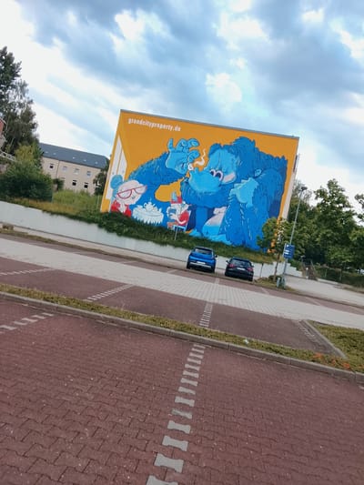 The image depicts an exterior view of a building featuring a large mural. The mural, painted on a yellow background, displays a blue, cartoonish monster-like character interacting with two smaller, human-like figures. The character on the right appears to be smoking. Text on the mural reads "grandcityproperty.de".
In the foreground, a paved parking lot is visible, laid with red and grey interlocking bricks. Two cars are parked in the lot: a blue hatchback on the left and a black hatchback on the right. A blue parking sign with a white 'P' is mounted on a pole to the right of the cars.
Behind the parking lot and the mural, there is a building with multiple windows and a grey roof. Trees and greenery are present along the side of the building and surrounding the area. The sky above is overcast with a mix of white and grey clouds.
Based on the "grandcityproperty.de" text on the mural, the location is likely associated with Grand City Property, a real estate company. While this specific image doesn't directly provide a street address, Grand City Property operates in multiple cities in Germany.
A forensic examination of the image reveals no discernible evidence of crime or distress. The scene is calm and ordinary. The lighting suggests it is daytime, possibly late afternoon or early evening given the cloud cover. The composition of the image, with its low angle and tilted perspective, suggests it was taken by a person standing in the parking lot.
Further research identifies this mural as being located in Chemnitz, Germany, specifically on the facade of a building managed by Grand City Property. The mural is part of a larger art project aimed at revitalizing urban spaces.