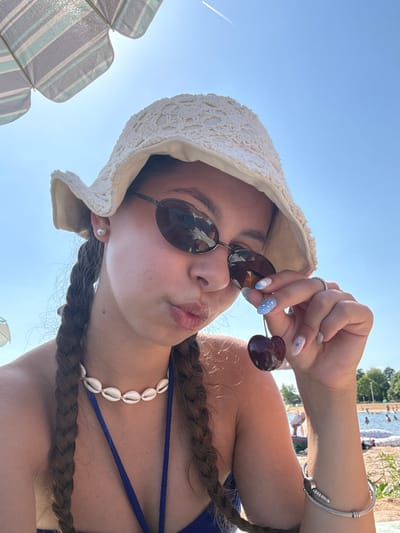 A young woman with braided hair and a white bucket hat is shown in a close-up, self-taken photo. She is wearing sunglasses and a blue bikini top, adorned with a seashell necklace. The woman is holding a cherry to her lips, and her nails are decorated with blue and white patterns. She appears to be at an outdoor swimming area, with a beach and people swimming in the background. The sky is clear and blue, and a portion of a striped umbrella is visible in the upper left corner. The overall scene suggests a sunny day at a recreational lake or beach. This location is identified as the Kinzigsee in Langenselbold, Germany.