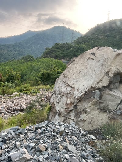 The image depicts a mountainous landscape near Aê Thi, Vietnam. In the foreground, there is a large, light-colored rock formation, with a pile of smaller, grey rocks and some vegetation at its base. Beyond the rock formation, there are layers of mountains covered with green trees and vegetation. Power lines and towers are visible on one of the mountains in the background. The sky is overcast and cloudy, suggesting it might be early morning or late afternoon. The angle of the image indicates it was taken from a hillside or elevated position.