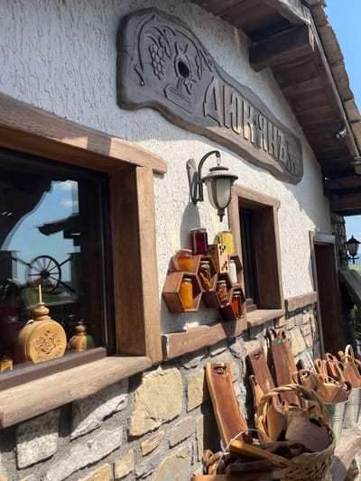 This image presents a low-angle view of the exterior of a building, likely a shop, on a bright, sunny day. The scene is characterized by rustic and handcrafted elements.
The building features an off-white, textured stucco wall, contrasted with dark wooden beams and window frames. To the right, a section of the wall is constructed from irregular, light-colored stone blocks.
A large, vertically oriented wooden sign is prominently displayed on the left stucco wall. It is intricately carved with Cyrillic text that reads "МЕДАРКА" (MEDARKA), and features decorative motifs, including what appears to be a stylized fish or bird at the bottom. Below this sign, a decorative wall lantern with a frosted glass shade is affixed.
Further right, several hexagonal wooden shelves are mounted on the wall, holding an assortment of glass jars. These jars contain various substances, likely honey or preserves, identifiable by their different colors and inclusions such as nuts or spices. Below these displays are two windows with robust dark wooden frames. The lower window's glass reflects a clear blue sky and objects inside the building, notably a red-spoked wagon wheel and a carved wooden cylinder featuring a rose design and partial text that might read "ARIA".
On the far right, against the stone wall, an array of wooden crafts is displayed, including several dark wooden cutting boards of various shapes, some with handles, and woven baskets. These items appear to be merchandise.
The bright sunlight casts distinct shadows, indicating clear weather. The overall impression is one of traditional craftsmanship and natural products, fitting a rural or artisanal setting in Medovo, Bulgaria. No people are visible.