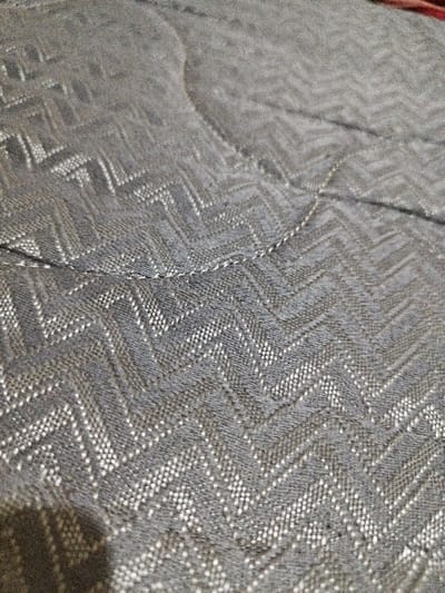 A close-up photograph captures a gray textile surface with a distinct zig-zag (chevron) pattern. The pattern is formed by alternating lighter gray or white threads against a darker gray woven background, creating a textured appearance. A single, visible line of curved machine stitching traverses the upper-mid section of the fabric. No individuals, specific objects other than the fabric, actions, or interactions are present. The environment is an indeterminate indoor setting. This scene is located in Situhiang, Indonesia.