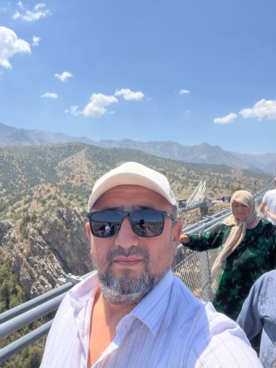 The image depicts a selfie taken on a suspension bridge in the vicinity of Dardar, Uzbekistan. The foreground is dominated by the face of a man wearing sunglasses and a white baseball cap, with a light-colored collared shirt. His reflection in the sunglasses shows a few other people. To his right, further along the bridge, a woman in a green patterned dress and a light-colored headscarf is visible. Another person in a grey, ornate garment is partially seen on the right edge of the frame. The bridge itself is a prominent feature, with metal railings and cables. In the background, a vast mountainous landscape stretches out under a bright blue sky with scattered white clouds. The mountains are covered in sparse vegetation. The overall scene suggests a tourist location.