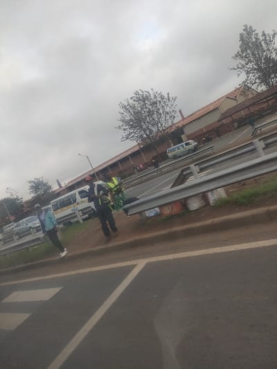 This image depicts a roadside scene in Gatongora ward, Kenya, under an overcast sky. The foreground shows an asphalt road with white solid and dashed lane markings, viewed from a slightly elevated position, possibly from within a vehicle. A metal guardrail separates the immediate foreground from a dirt shoulder and an adjacent road.
On the dirt shoulder, to the left of the guardrail, a group of at least four individuals is present. One person on the far left wears a light blue/green jacket and dark pants. To their right, another individual wears dark clothing with a red cap and a light-colored harness or vest. Next to them, a person wears a bright green or yellow safety vest over a patterned garment. Another individual is partially obscured behind the person in the green vest. Several white and red/orange bags are placed under the guardrail on the dirt shoulder.
Beyond the guardrail, on an adjacent road, two white minibuses (matatus) are parked or stopped. The closer minibus displays blue lettering and numbers, including "BOG" and "20." A silver-colored car is partially visible behind it. Further down the road to the right, another white minibus with blue and yellow markings is visible. In the background, a series of light-colored buildings with reddish-brown sloped roofs line the far side of the road. Trees with sparse foliage are scattered throughout the background. A street light pole is visible between the foreground and background activity.