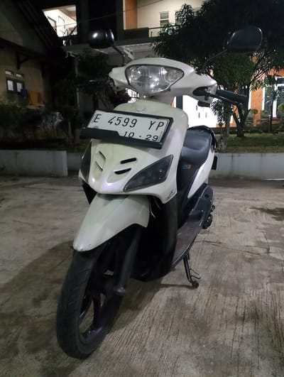A white scooter is parked on a concrete surface at dusk. The scooter is positioned facing forward, slightly angled to the left. A license plate with the characters "E 4599 YP" and the date "10.29" is attached to the front of the scooter. In the background, there is a building with windows and trees. The scene appears to be in the city of Cigadung, Indonesia.