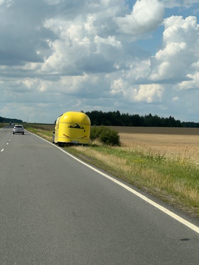 On a two-lane asphalt road, a yellow trailer with a mountain graphic and a license plate reading "A 9330 I-5" is parked on the shoulder. A white SUV is driving in the left lane ahead of the trailer. The road is bordered by a grassy shoulder and a field of dry wheat, with a dark green forest in the distance under a sky with large, white, puffy clouds. The scene appears to be in a rural area, possibly near Чырвонаслабодскі сельскі Савет, Belarus, given the context.