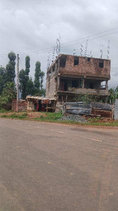 The image displays a scene in Moticho ward, Kenya, featuring a three-story building under construction, a makeshift kiosk, and a paved road under an overcast sky.
**Objects:**
* **Building:** A multi-story concrete and brick structure, primarily unfinished. The ground and first floors exhibit exposed concrete columns and slabs, while the second floor shows unplastered reddish-brown brick walls with open window apertures. Vertical rebar rods protrude from the roofline, indicating ongoing or planned construction.
* **Kiosk:** To the left of the main building, a small, weathered kiosk constructed from corrugated metal sheets and wooden planks is present. Its exterior features faded lettering and graphics.
* **Fence/Barrier:** A fence composed of vertically oriented corrugated metal sheets and horizontal wooden planks runs in front of the construction site and kiosk.
* **Construction Materials:** Piles of grey aggregate (likely gravel or crushed stone) are visible on the ground adjacent to the fence, near the building.
* **Road:** A dark, paved road with faint, discontinuous white dashed lines occupies the foreground, stretching from bottom-center towards the middle distance.
* **Vegetation:** Green trees and shrubs are dense in the background on the left. Sparse green foliage, including grass and weeds, grows along the roadside and around the structures.
* **Utility Pole:** A tall, slender wooden utility pole stands on the left side, behind the kiosk, with visible overhead wires extending across the frame.
**People:**
* Within the shaded interior of the makeshift kiosk, three indistinct figures are observable. Their specific actions or interactions are not discernible due to distance and resolution.
**Environment:**
* The sky is uniformly grey and overcast, suggesting a cloudy day.
* The overall scene conveys an impression of ongoing development and local commerce in a semi-urban or rural setting.