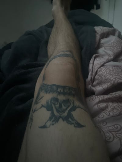 The image shows a close-up of a person's leg, with a tattoo on the knee area. The tattoo appears to be a dark, intricate design, possibly depicting an animal or a mythical creature. The leg is hairy and the skin tone is light. The background consists of dark blankets and a patterned fabric, suggesting the person is likely in a bedroom or on a bed. The lighting is dim, creating shadows and an intimate atmosphere. The image is taken from a slightly elevated angle, looking down at the leg. There are no other people visible in the image. The scene does not contain any recognizable landmarks or contextual clues that would place it specifically in the city of Paczkowo, Poland.