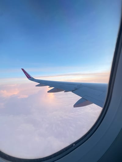 The image shows a view from an airplane window. The wing of the airplane is visible, with "wizzair.com" printed on the winglet. The sky is visible outside the window, with a gradient from blue to orange and pink near the horizon. The scene is likely at a high altitude. There is no visible information indicating that this image was taken in Okorsh, Bulgaria. There are no people or interactions visible. The environment consists of the inside of the plane and the external sky.