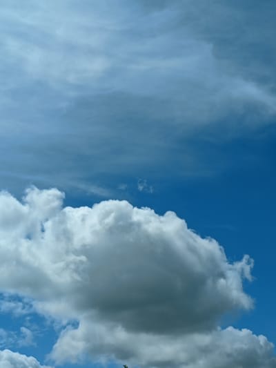 The image captures a dynamic sky with varying cloud formations and a bright blue expanse. In the foreground, large, fluffy cumulus clouds dominate the lower half of the frame, appearing dense and white, indicative of fair weather. Above these, thinner, wispy clouds stretch horizontally across the sky, painted in softer shades of gray and white, suggesting higher altitude atmospheric conditions. The overall lighting suggests it is daytime, possibly mid-afternoon, with a clear and vibrant blue sky peeking through the cloud layers. No people, structures, or specific landmarks of Peetri alevik, Estonia are visible in this image, which focuses solely on the atmospheric elements.
