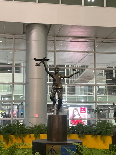 The image shows a bronze statue of a young boy holding a model airplane, situated in what appears to be an indoor public space, possibly an airport or a large transit hub. The statue is dark brown and depicts the boy in mid-leap, with his arms outstretched. He is wearing a short-sleeved shirt and shorts. In his right hand, he holds a larger model airplane, and in his left hand, a smaller one. The statue stands on a dark, cylindrical pedestal, which is in turn placed on a circular, dark base. Behind the statue, a large, gray, cylindrical column is visible, likely a structural element of the building. The background consists of a large glass window pane with multiple horizontal and vertical dividers, revealing reflections of the interior and possibly other parts of the building. Through the glass, one can discern interior lighting fixtures on the ceiling and glimpses of what might be shops or information displays. There are also large, bright advertisements visible through the glass. One prominent advertisement features a woman wearing jewelry and a red floral dress, with the "KALYAN JEWELLERS" logo clearly visible. Green foliage, possibly potted plants or shrubs, is arranged in front of the base of the statue and along a bright yellow planter box, adding a touch of nature to the man-made environment. The overall impression is of a modern, well-lit public space with artistic elements. The reference to Raipur Khurd, India suggests this location is within that region.