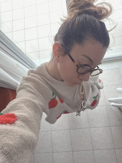 A forensic examination of the image reveals an indoor scene within the Municipal Unit of Corinth, Greece. The primary subject is a female individual, photographed from an overhead angle, capturing her head and upper torso. Her dark brown hair is styled in a messy bun. She wears eyeglasses with dark, tortoise-shell frames and a thin, gold-colored chain necklace. Her attire consists of a light-colored, possibly cream or beige, knitted sweater or robe adorned with multiple red strawberry motifs. The subject's gaze is directed downwards. The visible environment includes a light-tiled floor composed of square tiles. To the left, a white curtain or panel, possibly a shower curtain, is partially visible, with an indistinct reddish-orange object positioned behind it. To the right, a white, textured object, consistent with a folded towel or a ceramic fixture, is partially observed.