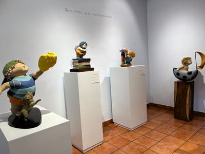 An art exhibit features four child-like sculptures displayed on pedestals. The first shows a child holding a large, yellow form above a dog. Another rests on a stack of books. The third appears colorful, with an attached, smaller figure. The last sits inside a dark, polka-dotted vessel with a sail, perched on a wooden pedestal. The walls are white, with Spanish script above the sculptures. The floor is tiled in terracotta.