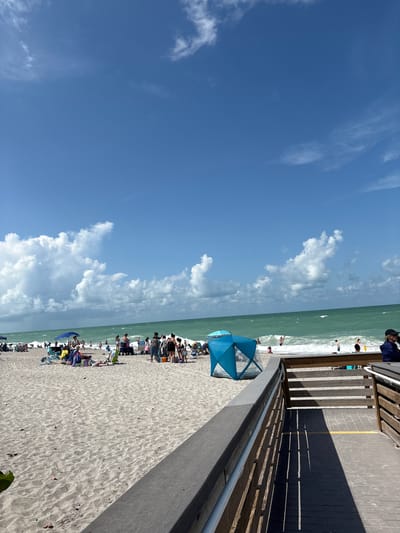 The image depicts a crowded beach scene on a sunny day. The sand is light in color, with numerous people scattered across it, many relaxing under umbrellas or in beach chairs. A turquoise ocean with moderate waves stretches across the horizon under a bright blue sky with scattered white clouds. To the right, a wooden boardwalk leads down to the beach, with railings and a yellow line marking a path. Several individuals are visible on the boardwalk and on the beach, some wading in the water or walking along the shoreline. A blue pop-up beach tent is set up on the sand. The overall impression is a vibrant and active recreational area.