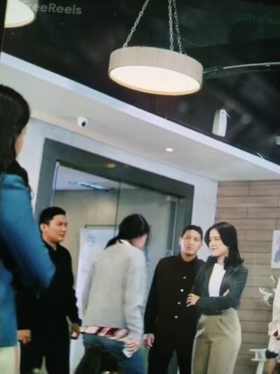 This media file captures a scene with several people indoors, possibly in an office or commercial building. In the foreground, a person wearing a blue jacket is partially visible on the left. Behind them, a man in a black shirt is looking to the left. To the right of the man, a woman in a dark blazer over a white shirt and light-colored pants stands with her arms crossed, looking to her right with a slight smile. Behind her, another person in a black jacket is partially visible. A person in a grey sweater and jeans is in the center, facing away from the camera. The setting appears to be a modern interior with a suspended light fixture overhead and a security camera mounted on the ceiling. The overall lighting suggests it is daytime, and the mood seems casual or professional. No specific weather conditions are discernible. The scene does not offer explicit cues about its location within Palangka Raya, Indonesia, beyond the given context. Text is visible in the upper left corner of the image, reading "reeReels."
