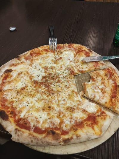 The image shows a pizza on a wooden board, on a dark brown table. A slice has been cut, and a fork and knife rest on the pizza and table. A bottle cap is also visible on the table. The pizza appears to be topped with cheese and tomato sauce, possibly in the city of Bayreuth, Germany.