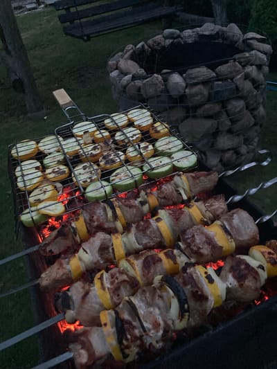 A close-up, eye-level shot captures a barbecue scene outdoors, likely in the rural setting of Матыкальскі сельскі Савет, Belarus. The foreground is dominated by several skewers of meat and vegetables cooking over glowing red embers. The meat appears to be pork, interspersed with yellow and white onion slices, and some darker charred pieces. Behind the skewers, a metal grilling basket holds sliced zucchini, some charred and others lightly grilled. In the background, a rustic stone fire pit is visible, with dark, empty space within its circular stone structure. To the left, a dark wooden swing bench is partially visible, and a rough-hewn wooden post stands nearby. The ground is covered in dark green grass, suggesting an evening setting.