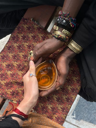 Two individuals are depicted holding a glass pitcher containing a yellowish liquid. One individual possesses darker skin and is adorned with multiple bracelets, while the other has lighter skin and wears a ring. The pitcher appears to be passed between them. They sit upon a bench covered with a patterned fabric, which appears to be placed on top of the bench. Based on the provided information and appearance of the individuals, it is likely that the photo was taken in the city of Boudha, Nepal, a suburb of Kathmandu, known for its Boudhanath Stupa.