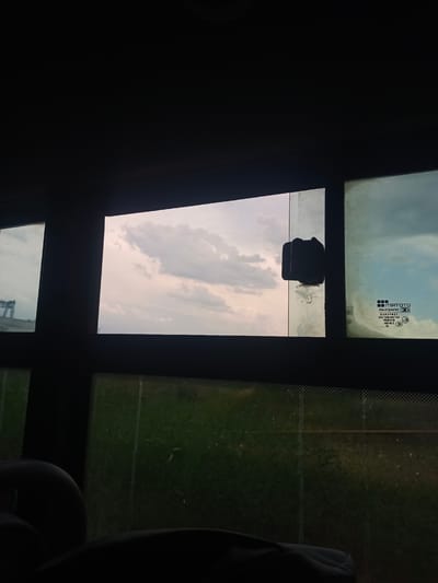 A view from inside a vehicle, looking out through windows at a cloudy sky and green landscape. In the central window, a black suction cup is attached to the glass. The window to the right has a logo and text printed on it, indicating it is tempered glass. In the foreground, dark shapes suggest the interior of the vehicle, possibly seats. The scene appears to be on the outskirts of Boralday, Kazakhstan, given the context provided.