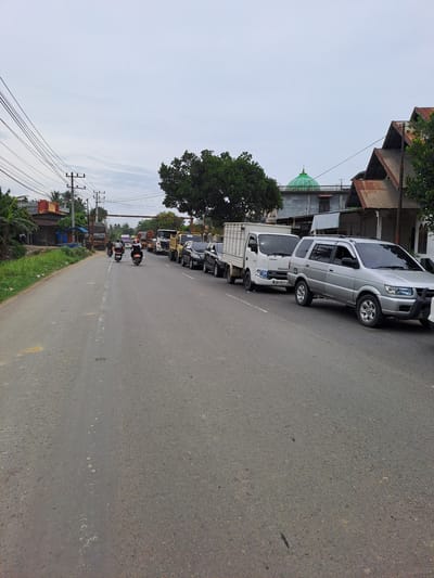 The image depicts a street scene in Kulu, Indonesia, characterized by moderate traffic congestion. A line of vehicles, including cars, trucks, and motorcycles, is stationary or moving slowly on a paved road. On the right side of the road, a silver SUV is prominently featured, followed by a white truck with a cargo box. Further back in the queue, other vehicles are visible, including a yellow dump truck and several passenger cars. On the left side of the road, two motorcycles are visible, one with a rider wearing a helmet and the other with a pillion rider. In the background, there are buildings with corrugated tin roofs and a green domed structure, likely a mosque. Trees and utility poles with numerous wires are also present, contributing to the urban and somewhat cluttered environment. The sky is overcast, suggesting a cloudy day. The road surface appears worn with some visible cracks and patches.