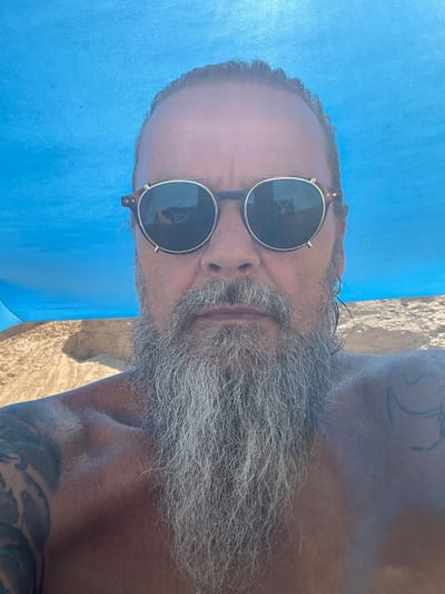 A man with a long grey beard and tattoos on his shoulder is looking at the camera while wearing sunglasses. He appears to be outdoors in a sandy area with a blue tarp above him, suggesting a beach or picnic setting. The image was taken in Ga'ash, Israel.