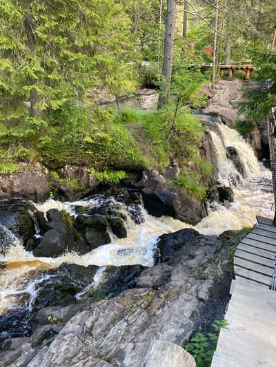 The image depicts a natural waterfall cascading over dark, rocky terrain in Ruskeala, Russia. The water is a muddy brown color, indicating possible sediment content. Lush green foliage, including trees and grass, surrounds the waterfall and clings to the rocks. In the background, a wooden bridge or walkway is visible, with two individuals standing on it, one wearing a red shirt and the other a camouflage-patterned outfit. The foreground features a wooden pathway with steps leading away from the waterfall. The scene suggests a scenic outdoor location, likely a tourist attraction.