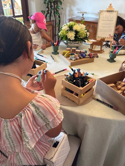 Here is a detailed description of the image:
**Scene**: The photo captures a well-lit, indoor scene where a group of people are engaged in an arts and crafts activity. The setting appears to be a casual event, possibly a child's birthday party or similar gathering.
**Location**: There's no definite evidence within the image indicating the city. However, the request indicates that the location is La Molina, Peru.
**People**:
* Several individuals are visible, including a child wearing a pink New York Yankees cap, and an adult who is wearing a white dress with red stripes and small green plants printed on it. They are sitting around a table and focused on crafting activities.
**Subjects**:
* The primary subject of the activity seems to be decorating wooden toy cars or blocks. There are various colored markers and crayons placed on the table, suggesting this is the medium used for decoration.
**Objects**:
* A wooden crate filled with colored markers is present on the table, alongside a collection of wooden toy cars/blocks.
* Other objects include an easel displaying a sign that reads "BABY CAR EMILIO" with instructions on decorating a car for Emilio, as well as flower arrangements and decorative items.
* A white quilted handbag sits on the couch next to the woman who is wearing a white dress with red stripes and small green plants printed on it.
**Actions**:
* The people in the image are actively drawing and decorating the wooden toys, indicating an interactive and creative atmosphere.
**Interactions**:
* The individuals seem to be engaged in collaborative or shared activity, as evidenced by their close proximity and focused attention on the crafting tasks.
**Environment**:
* The environment appears to be a well-furnished interior. The lighting is bright, creating a cheerful and inviting atmosphere. A large window to the left provides natural light, contributing to the overall brightness.