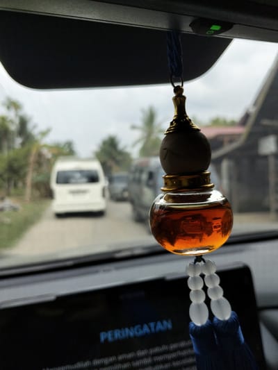 This is a close-up, indoor shot from inside a vehicle, looking forward. The primary subject is an amber-colored liquid in a decorative, gourd-shaped glass bottle hanging from the rearview mirror. The bottle has a gold-colored cap and base, and is adorned with a string of white beads and a dark blue tassel. In the background, the scene is slightly out of focus, showing a road with two white vans, and possibly other vehicles. Trees and buildings are visible on either side of the road, suggesting a rural or semi-rural setting. The sky appears overcast, indicating a cloudy day.
The text "PERINGATAN" is visible on a dark screen in the foreground, likely a car's dashboard display. This translates to "WARNING" in Indonesian. The lower part of the screen shows more text, also in Indonesian, which appears to be a safety message related to driving. The overall impression is one of being a passenger in a vehicle, possibly in a tropical or subtropical region, given the vegetation. The image evokes a sense of everyday travel. The lighting suggests it is daytime.