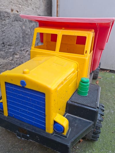 A close-up, eye-level shot focuses on a yellow, red, and blue plastic toy dump truck parked on a mottled gray and green surface, possibly concrete or asphalt. The truck's front features a blue grille and a yellow hood with a blue circular emblem and a yellow cap. A green barrel-shaped cap sits on the truck's black step. The truck's red bed is raised slightly. The background consists of a textured gray wall with some discoloration. The scene is outdoors, with scattered dry leaves on the ground. The lighting is natural, creating shadows and highlights on the truck's plastic surfaces. No people are present. The image does not provide any specific information about the location within Gorkovskiy, Russia.